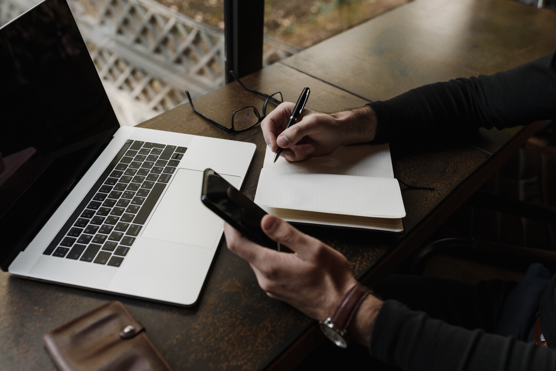 Hände einer Person. Sie hat Laptop, Smartphone, Stift und Papier vor sich.